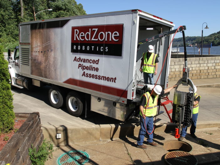Three workers lower a robotic crawler into a manhole for an Multi-Sensor inspection using a winch system attached to a RedZone Robotics box truck.